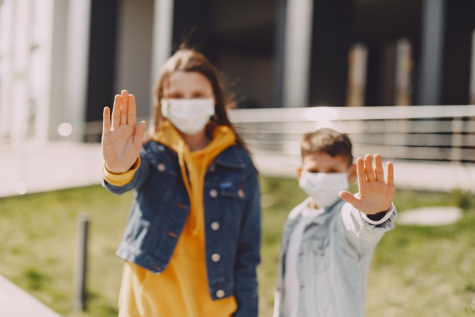 Young boy and girl in trendy casual clothes and medical masks standing on street and extending arms forward with raised palms while showing stop coronavirus