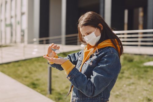 Free Side view of focused young girl with long dark hair in casual wear and medical mask pouring sanitizer on hands while standing on street near modern building Stock Photo
