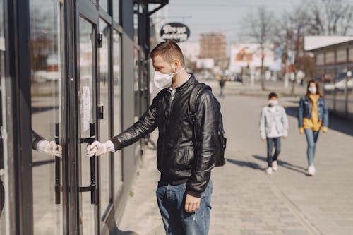 Side view of young guy in medical mask and disposable glove standing on sidewalk near shop and holding door handle while entering