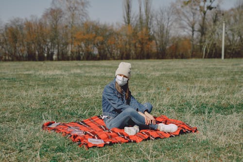 Free Young girl with medical mask and hat sitting with crossed legs on checkered blanket on grass and resting on lawn during weekend while having picnic Stock Photo