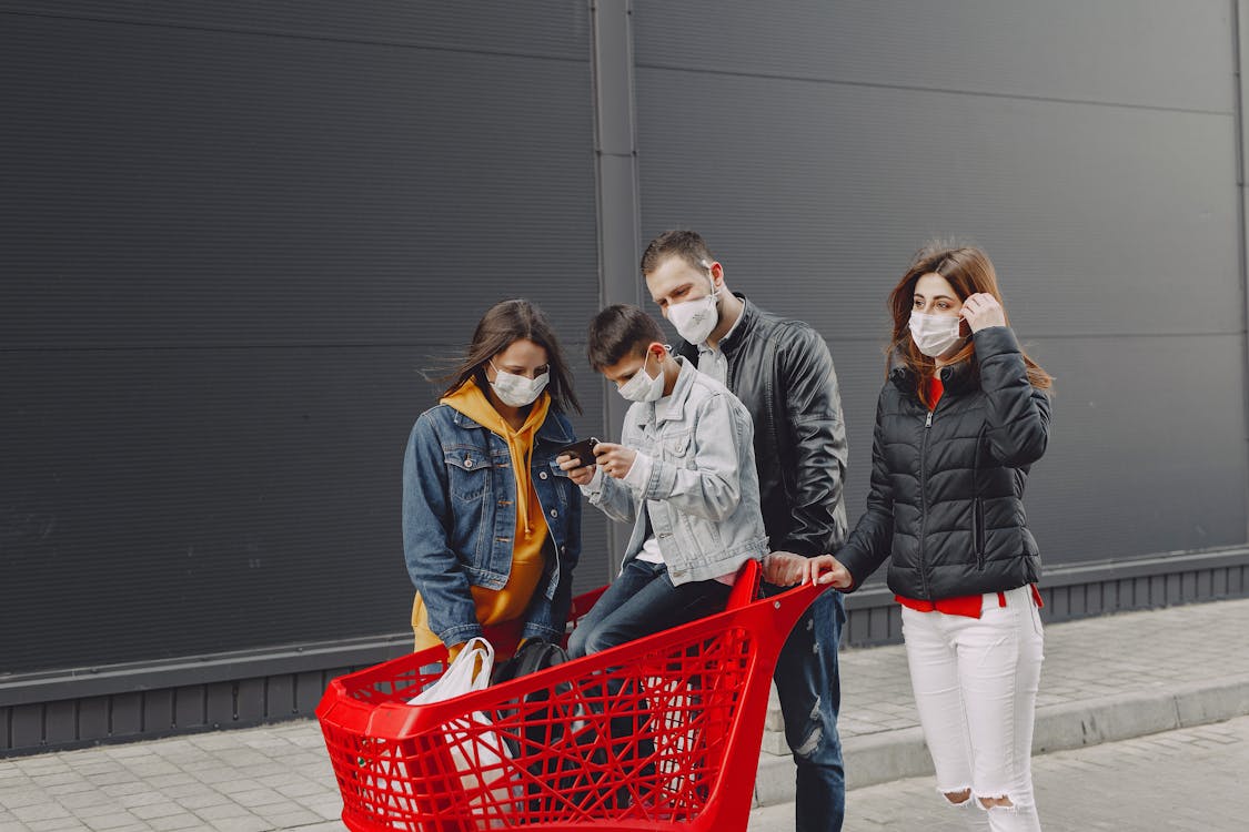 Free Happy family in casual clothes and medical masks standing on street after shopping while kid sitting in trolley and playing games on smartphone Stock Photo