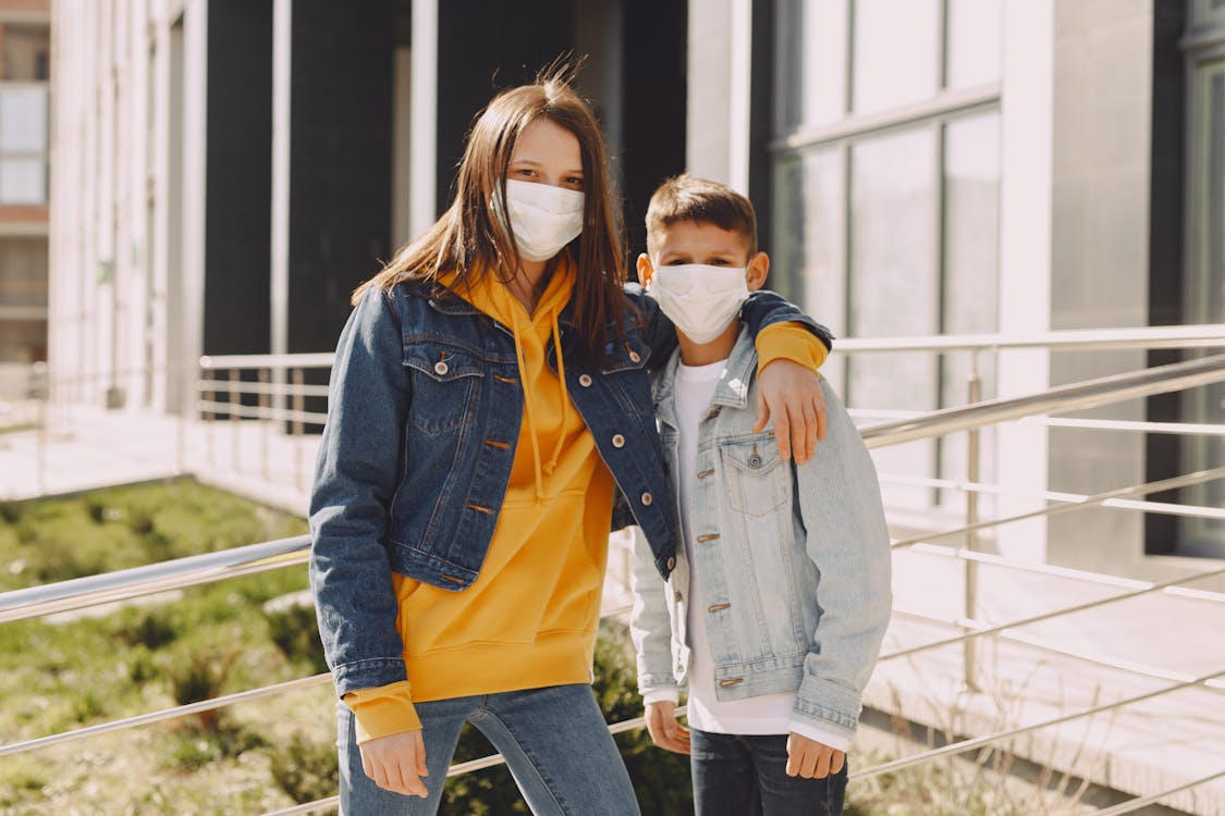 Free Children in casual clothes and protective masks standing on street near entrance of modern building while girl hugging boy in back lit Stock Photo