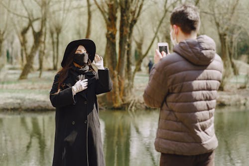 Young woman in warm outerwear and protective mask posing in park and adjusting hat while boyfriend in warm clothes and protective mask taking photo against river