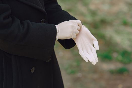 Side view crop unrecognizable female in black warm coat standing in park and putting on hands white protective medical gloves in daylight