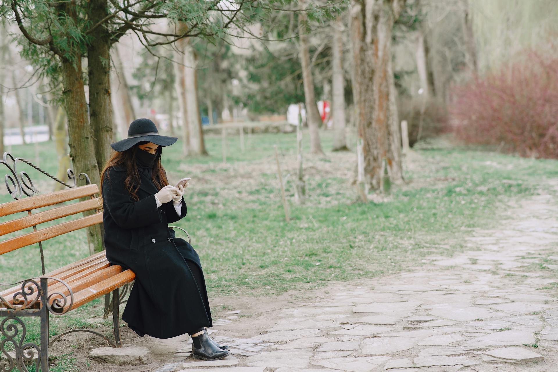 A woman in a park wearing protective clothing and mask, using her smartphone on a bench during a spring day.