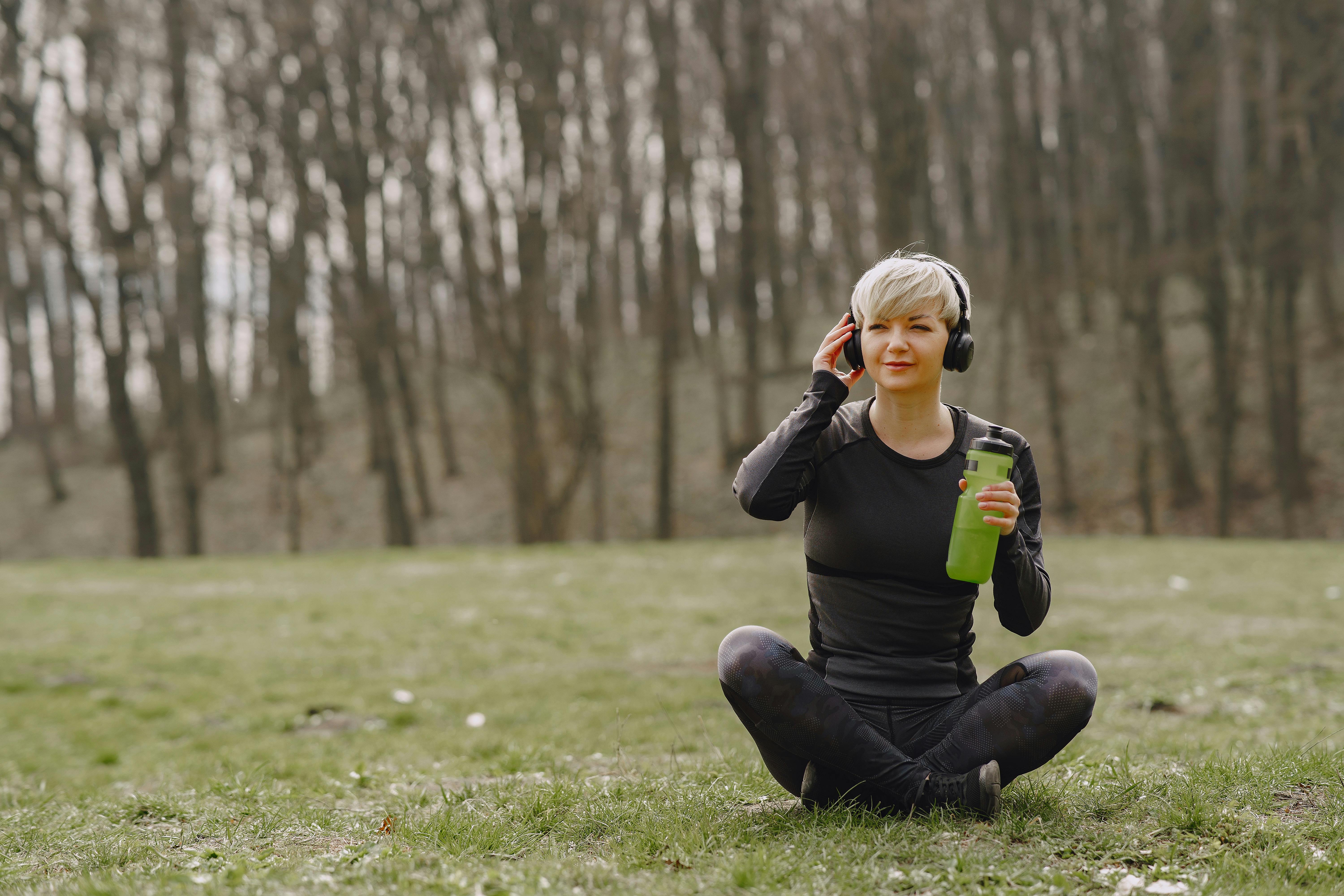 young fit woman in headphones sitting on ground holding bottle of water and listening to music