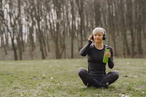 Young fit woman in headphones sitting on ground holding bottle of water and listening to music