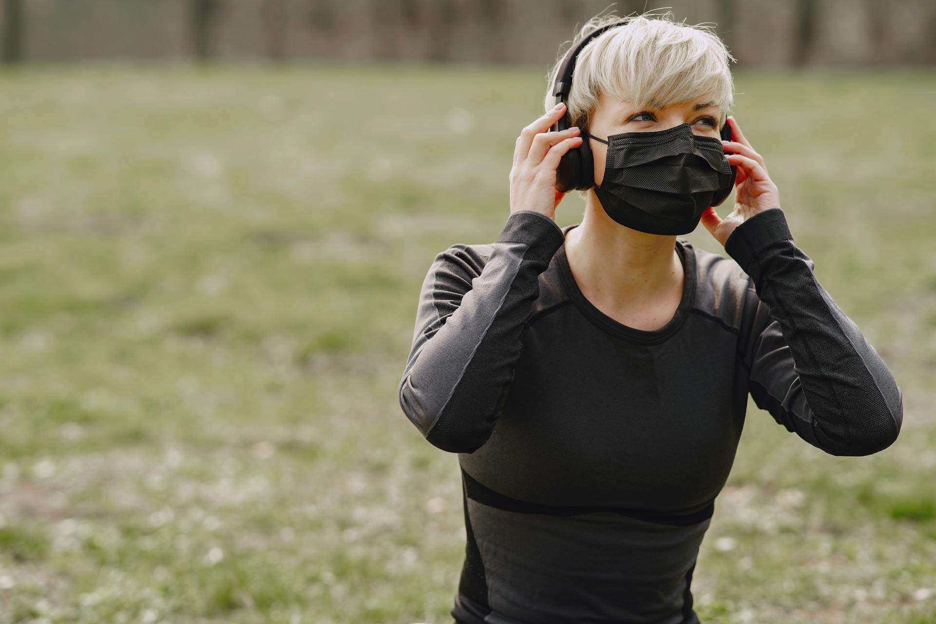 Young female in sportswear with black protective mask looking away and adjusting headphones while having rest after working out in park