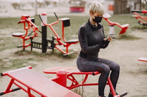 Serious young woman in face mask using smartphone sitting on street simulator