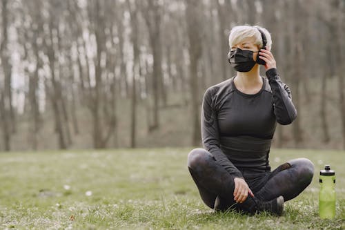 Free Full length of young fit female in sportswear and black protective mask with headphones and bottle of water sitting relaxed on ground of meadow near forest with legs crossed while listening to music and looking away Stock Photo
