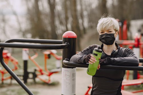 Stylish woman in medical mask with bottle of water on sports ground in street