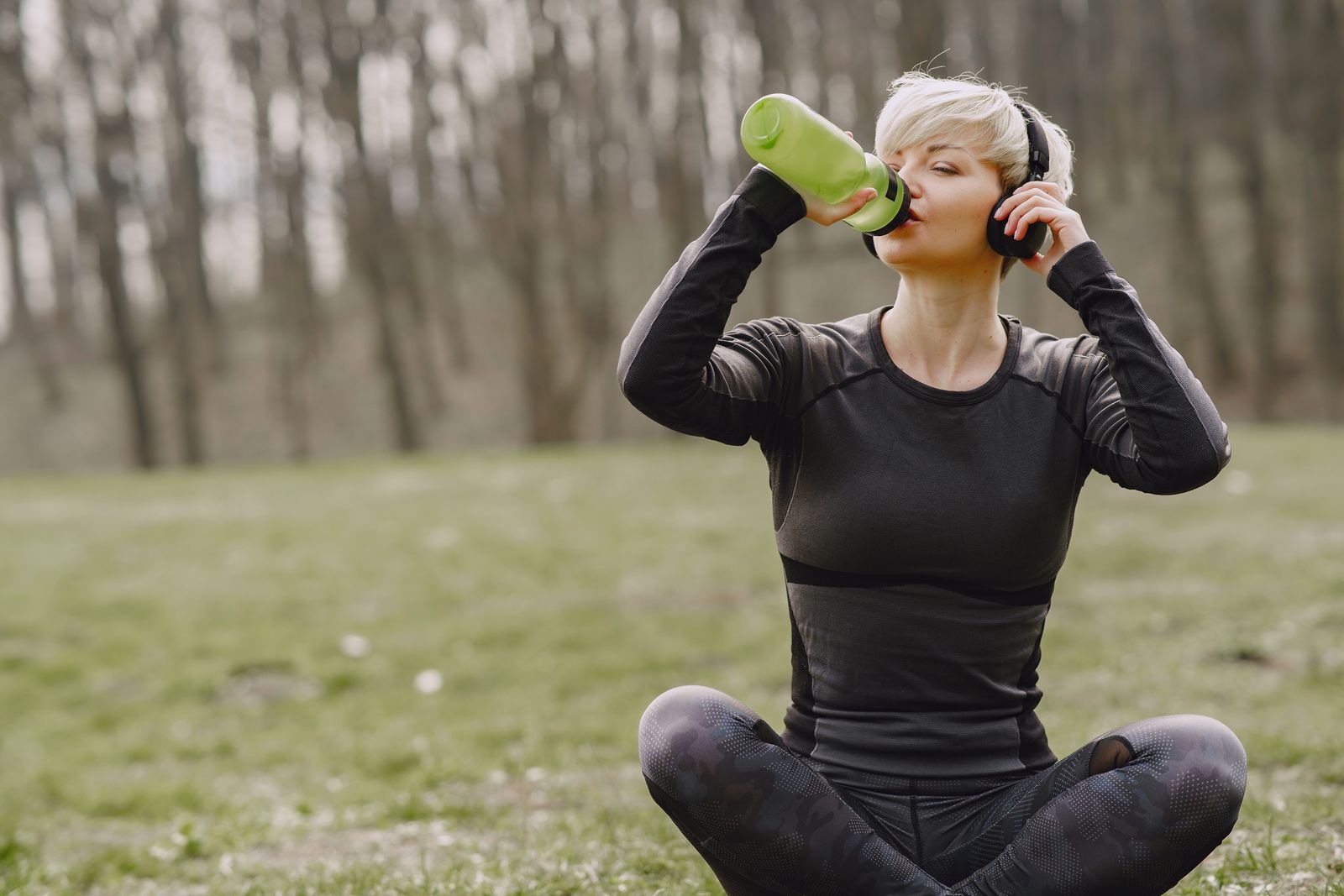 Woman in athletic wear sitting in lotus pose outdoors in a park holding a water bottle with eyes closed