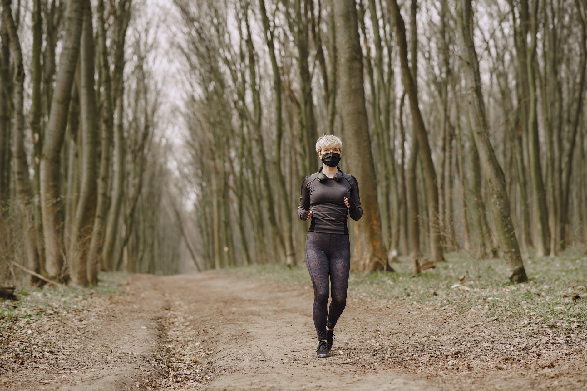 Full body woman in sportswear and disposable mask running along alley in park with headphones on neck during pandemic making workout