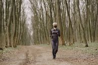 Full body woman in sportswear and disposable mask running along alley in park with headphones on neck during pandemic making workout