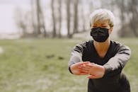 Strong female athlete in medical mask warming up before training in forest during pandemic