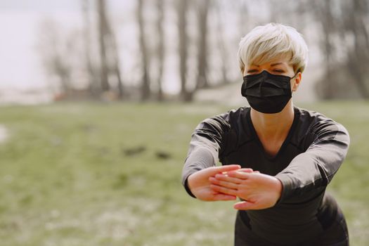 Strong female athlete in medical mask warming up before training in forest during pandemic