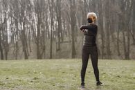 Full length young female in sportswear and mask making turns and kneading upper back while standing on green meadow in park