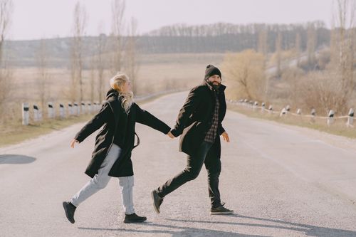 Free Side view full body of cheerful couple in casual warm outerwear holding hands and crossing road in rural area in spring morning Stock Photo