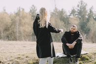 Blond girlfriend in back lit wearing black warm coat passing medical protective mask to boyfriend while spending time together in rural lawn during coronavirus outbreak