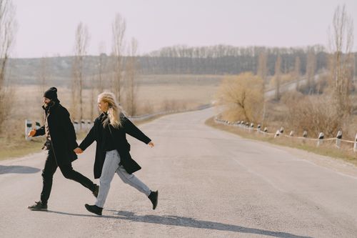Free Full length side view of couple in casual outerwear running across empty road and holding hands in rural area in spring morning Stock Photo