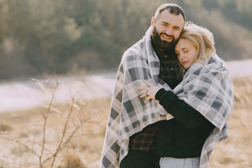 Free Cheerful loving cuddlers in back lit hugging while standing in warm casual clothes under cozy soft grey plaid in countryside during spring morning Stock Photo
