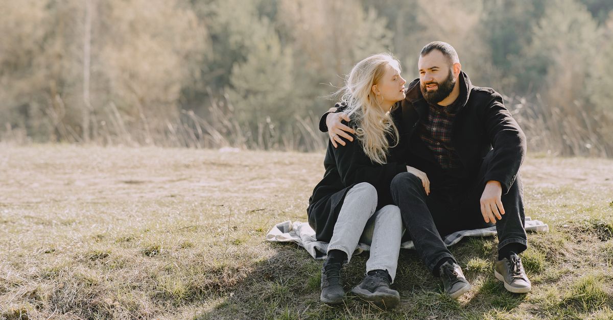 Positive dreamy couple in casual outerwear embracing each other and having rest together on grassy lawn while talking and spending time in countryside
