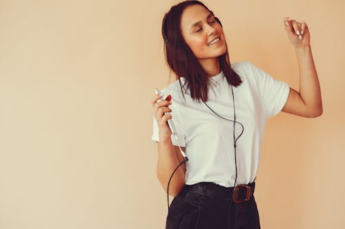 Happy young female in casual clothes smiling and dancing with eyes closed while listening to good music in headphones in studio on beige background