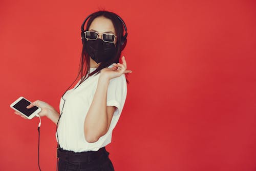 Side view of anonymous content stylish lady in sunglasses and protective mask using smartphone and listening to favorite music in headphones against red background