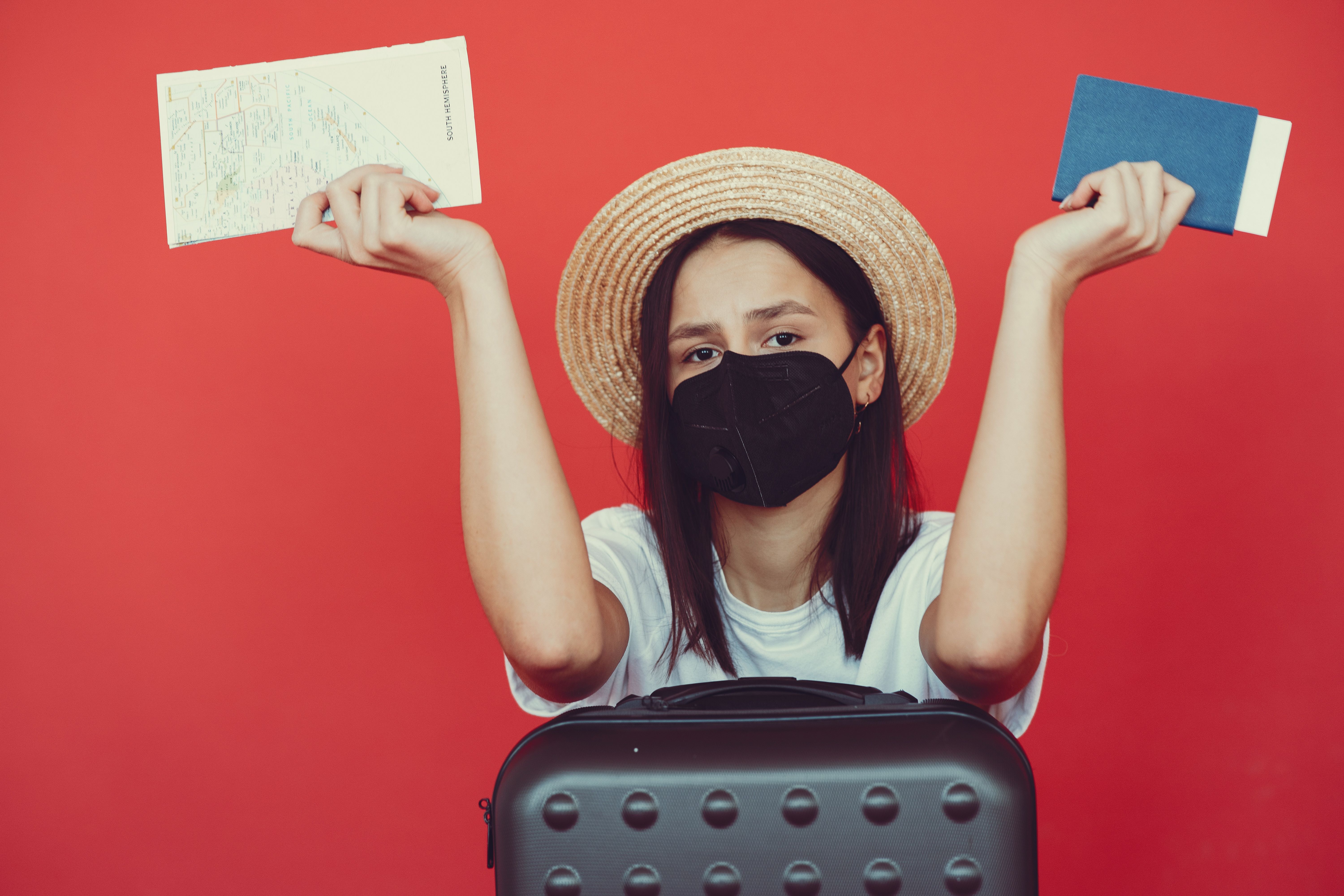 Traveller wearing a face mask holds a passport and map behind a suitcase