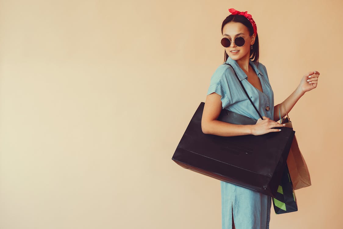 Free Side view of happy young shopaholic female wearing trendy blue dress and sunglasses carrying many shopping bags in studio on beige background Stock Photo