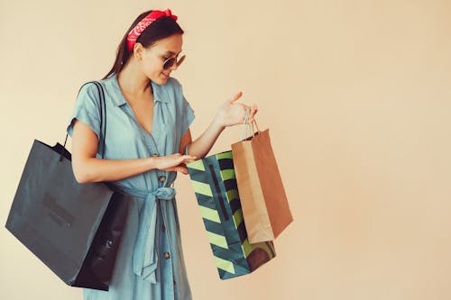 Free Cheerful young woman in trendy sunglasses and red headband looking into paper bags and holding shopping bag on shoulder during shopping on beige background Stock Photo