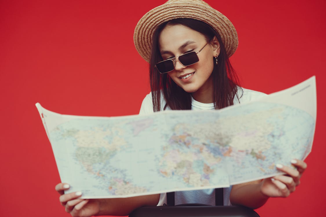 Free Smiling young woman in stylish hat and sunglasses choosing destination on paper map while standing with luggage on red background Stock Photo