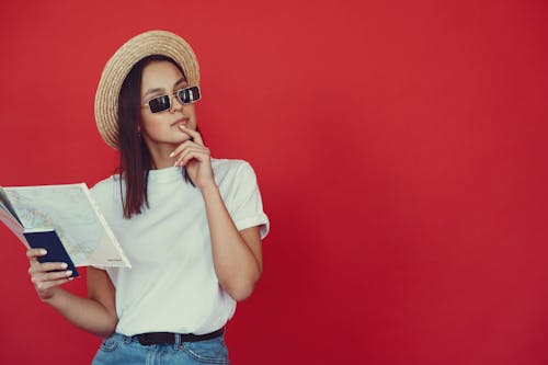 Thoughtful young woman in retro hat and trendy sunglasses holding map and touching chin while looking pensively away on red background