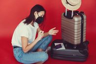Side view of young woman in casual clothes and medical mask sitting near luggage with passport and tickets while disinfecting hands on red background