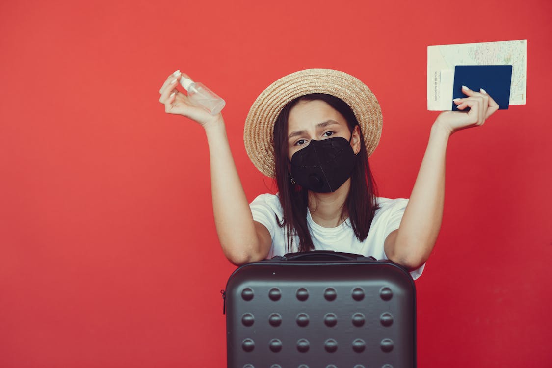 Free Young woman in medical mask and wicker hat holding passport and sanitizing spray while sitting behind suitcase and shrugging hand on red background Stock Photo