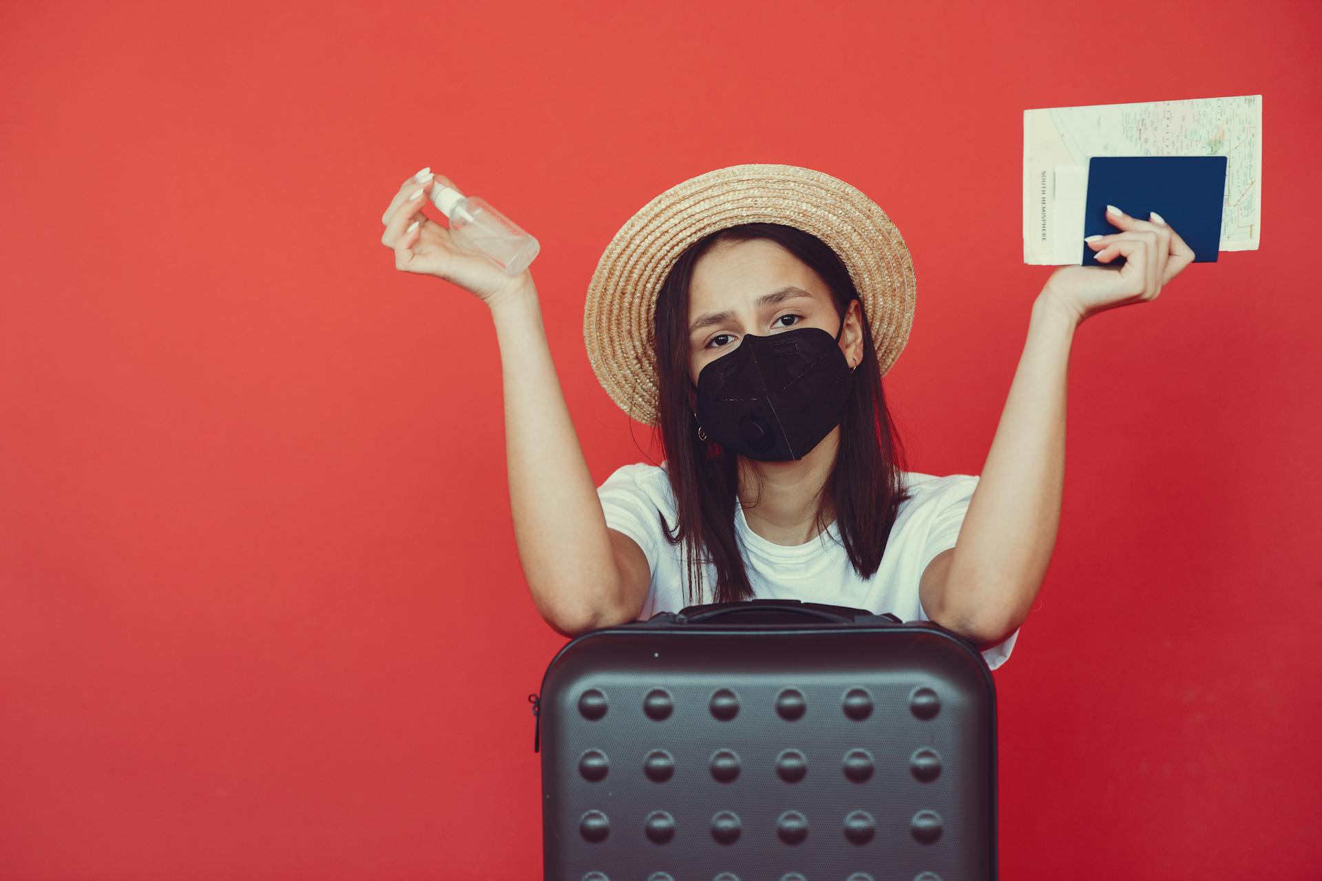 Young woman in medical mask and wicker hat holding passport and sanitizing spray while sitting behind suitcase and shrugging hand on red background