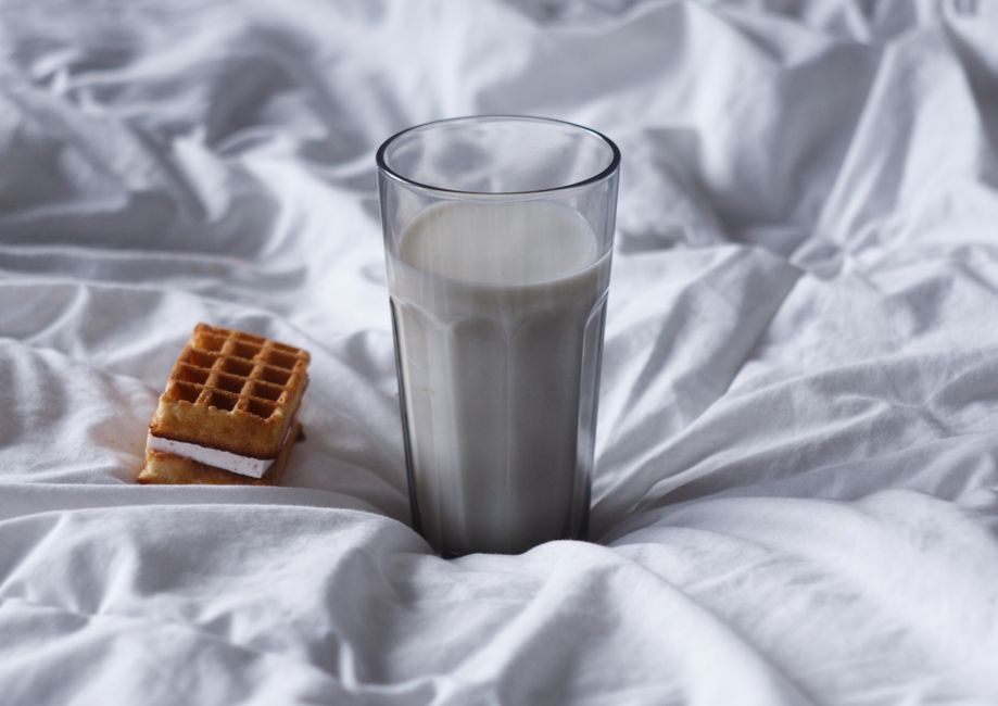 A glass of milk and a waffle resting on white bed sheets, providing a cozy breakfast scene.