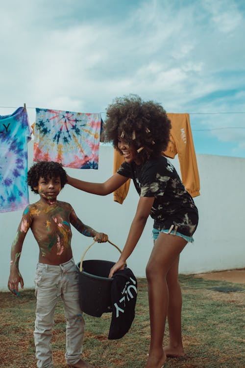 Full body of young smiling African American woman in casual clothing with fluffy hair stroking hair of shirtless black boy holding bucket and looking at camera
