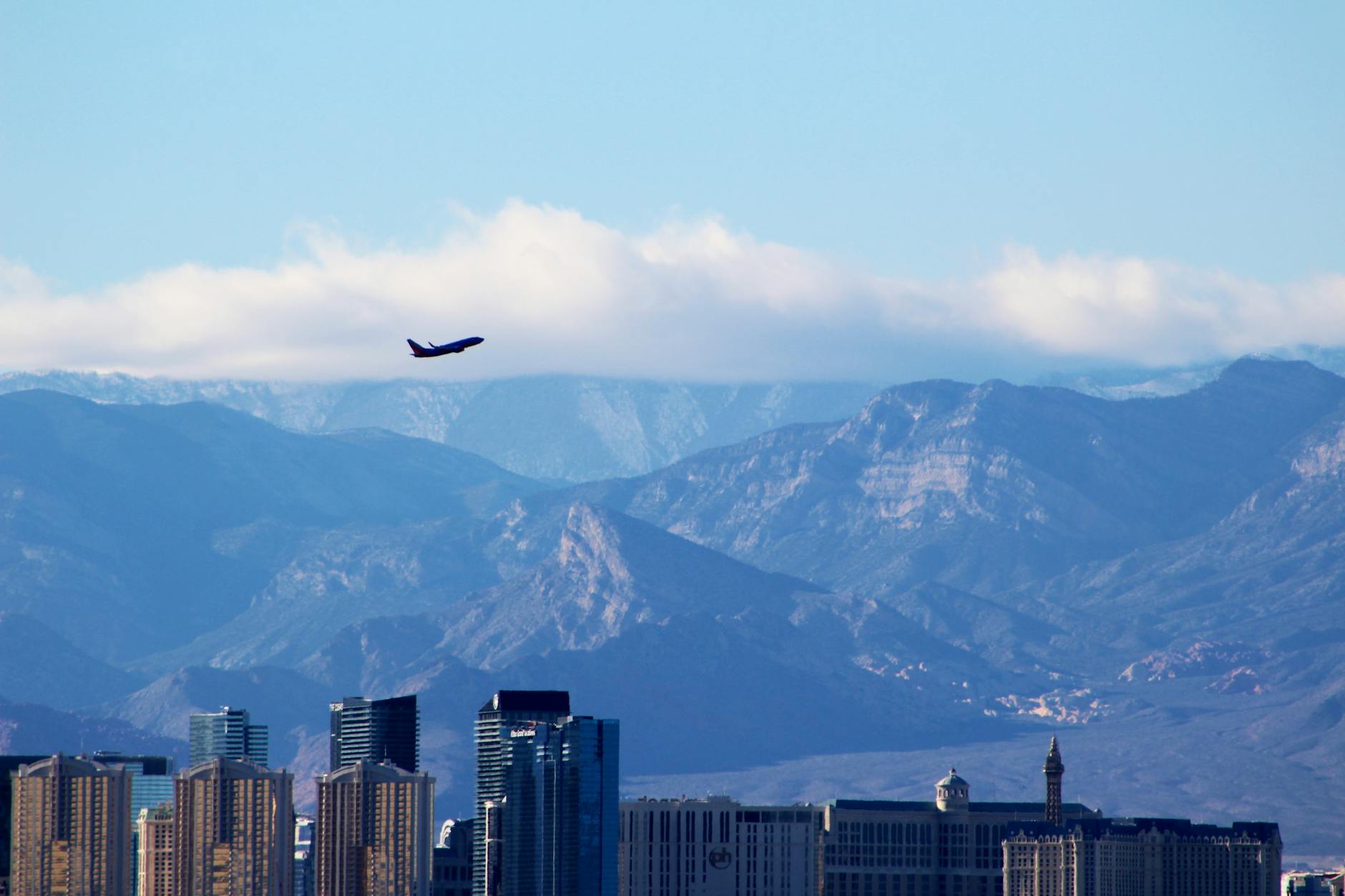 An airplane soars above the Las Vegas skyline with mountains and a cloudy sky in the background.