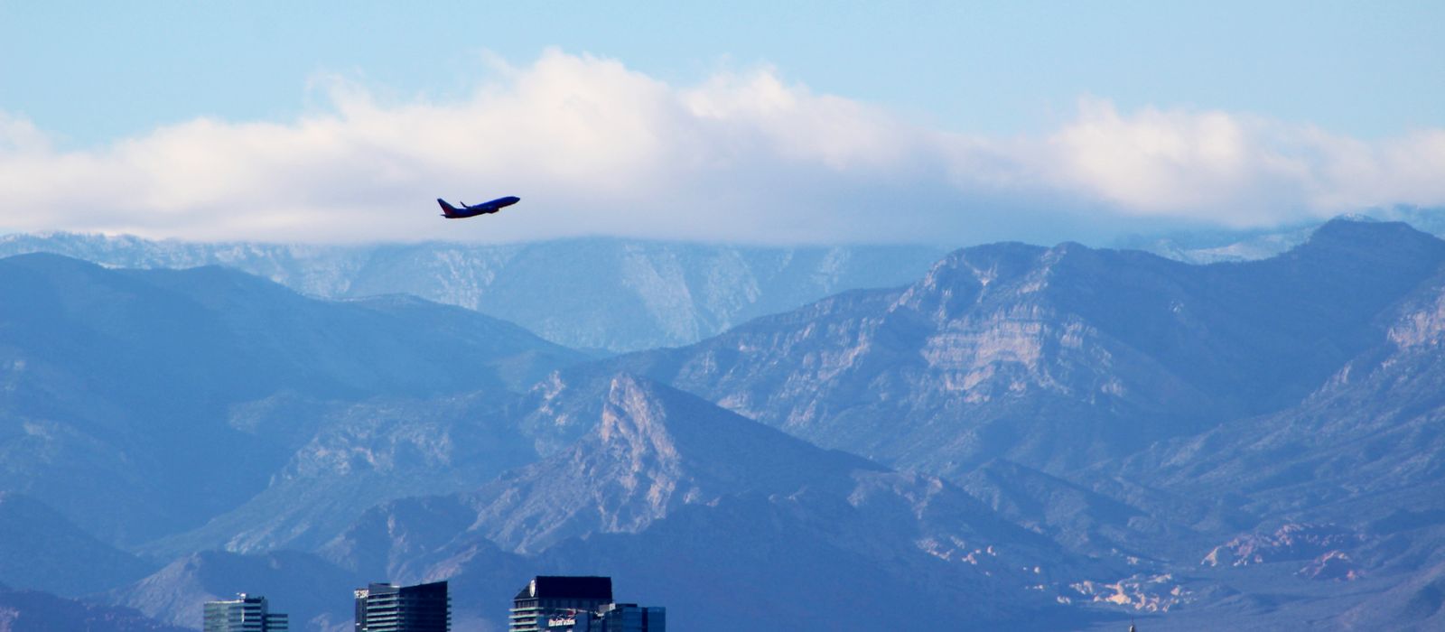 An airplane soars above the Las Vegas skyline with mountains and a cloudy sky in the background.