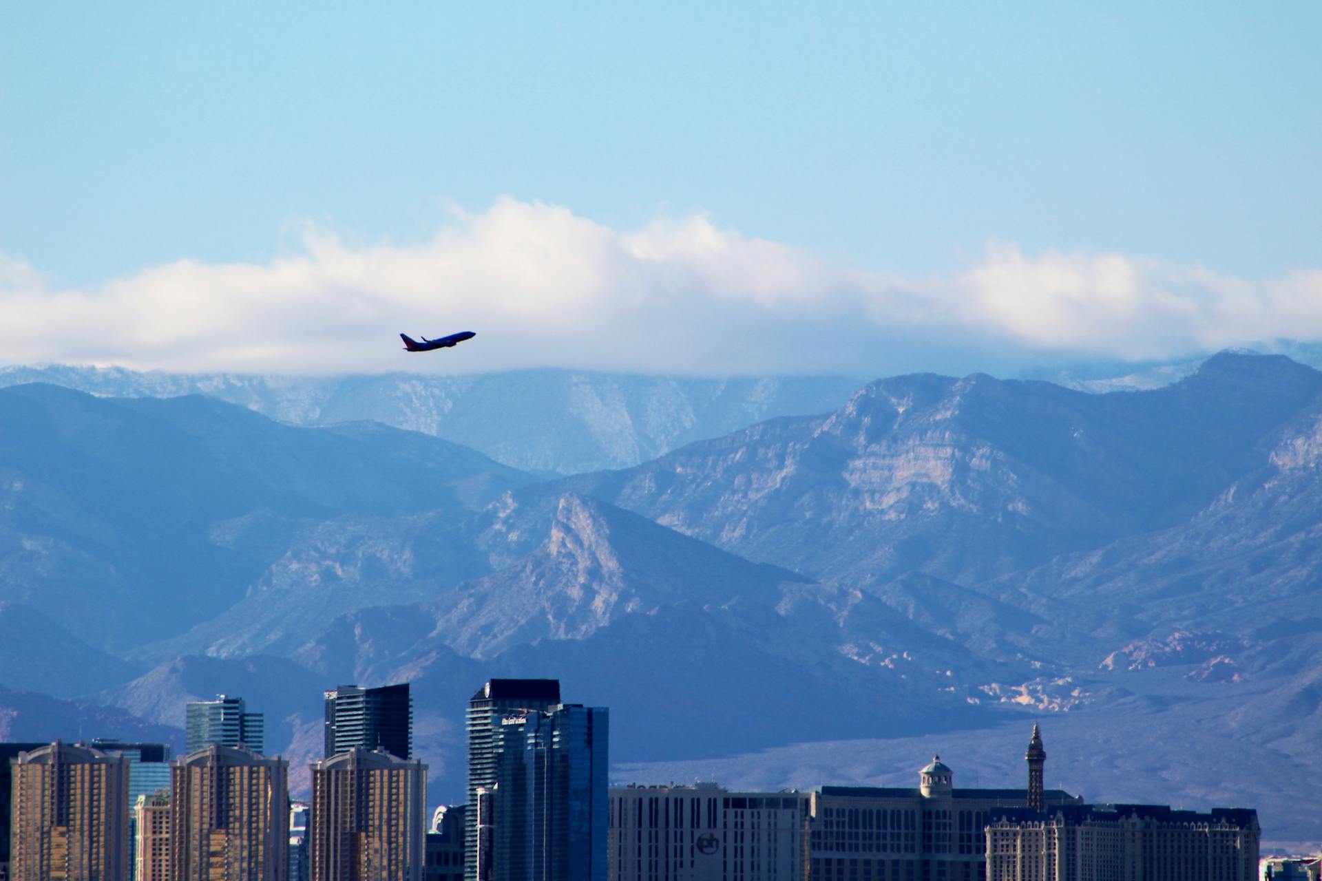 An airplane soars above the Las Vegas skyline with mountains and a cloudy sky in the background.