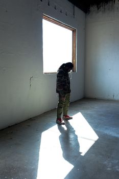 一个穿着温暖衣服、头发扎起来的无脸跟踪者，正站在阳光下旧破房子空房间的窗边，全身俯视
