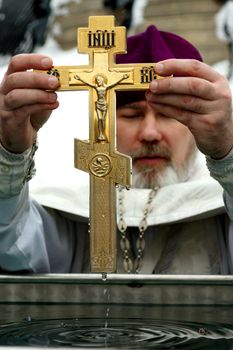 Concentrated priest in clothing for church ministry standing in front of water reservoir and holding wet Christian cross