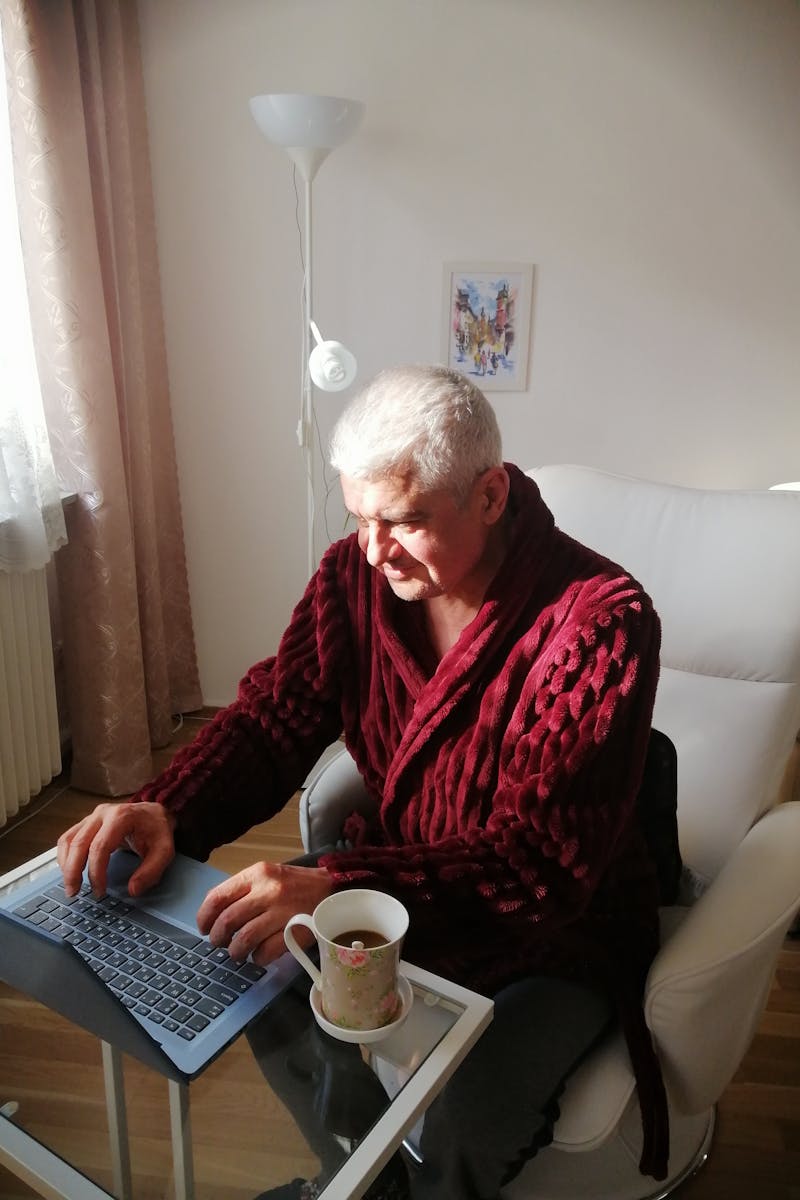 High angle of serious aged man in bathrobe having coffee while working remotely at home