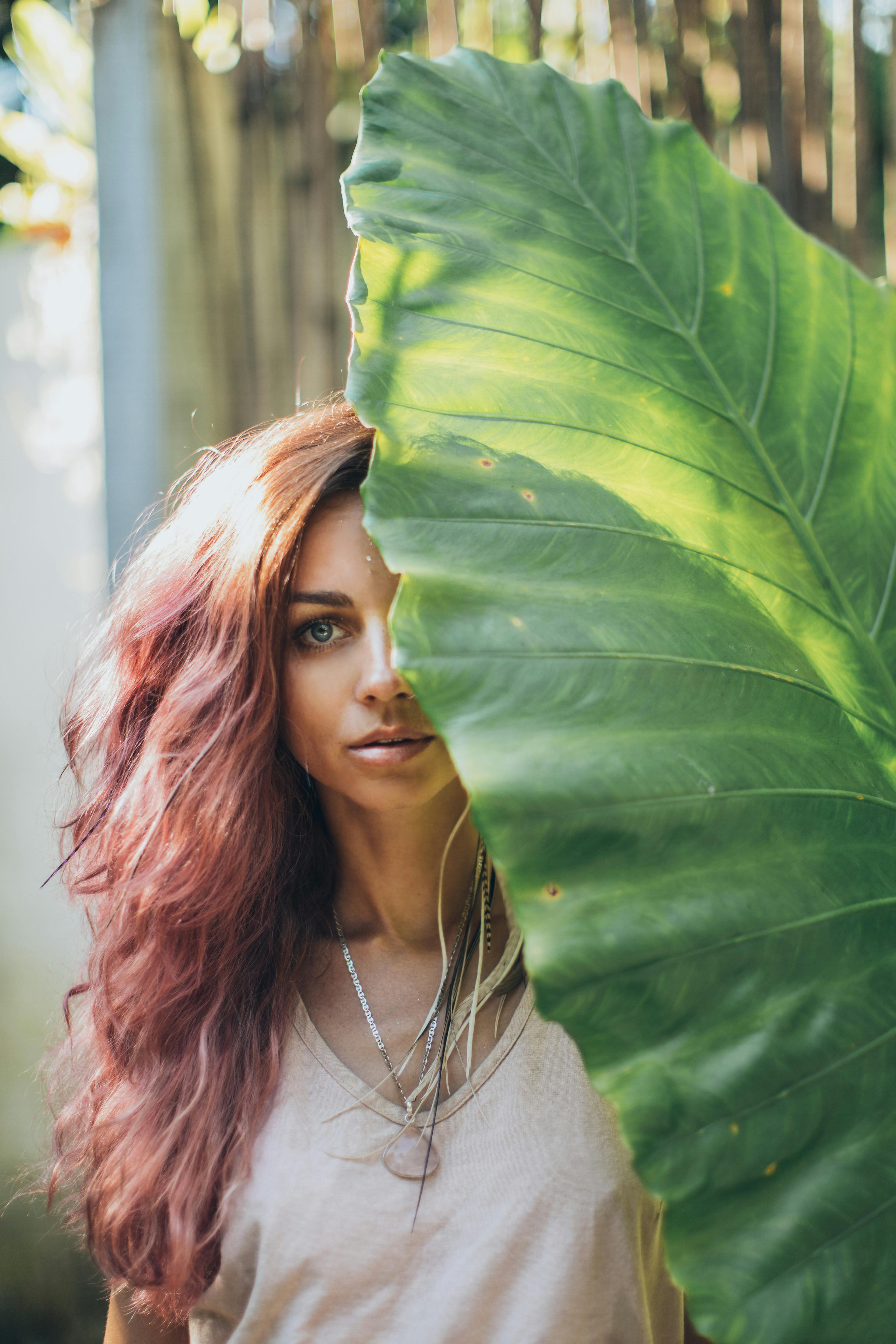 Young attractive female with pink hair and bright makeup covering face with big green leaf