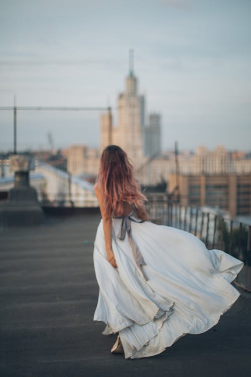 Back view of young female with long hair in white dress standing on rooftop against Moscow skyline