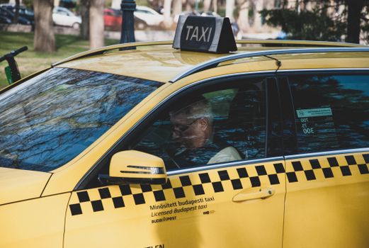 Gray haired man in glasses sitting on drivers seat of yellow taxi parked on street of Budapest