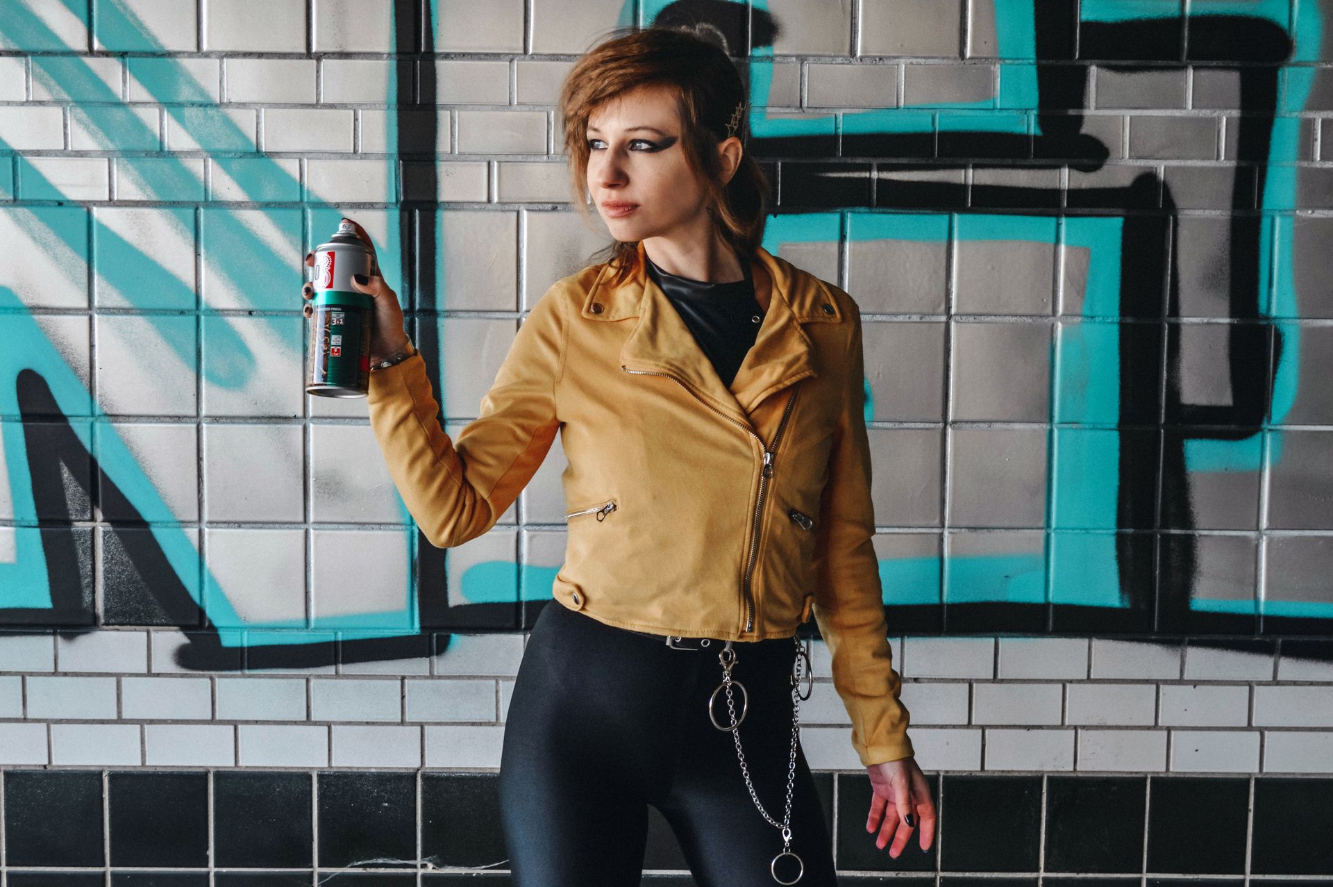 Confident young lady with stylish haircut in trendy clothes standing with spray paint near wall with graffiti on city street