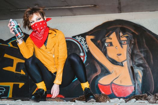 Young female in casual clothes with bandana on face resting near colorful graffiti on wall of modern building and holding spray paint