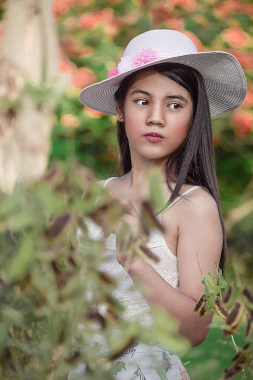 Charming young ethnic woman with long dark hair in stylish hat and summer dress standing among green bushes and looking away in summer garden at daytime